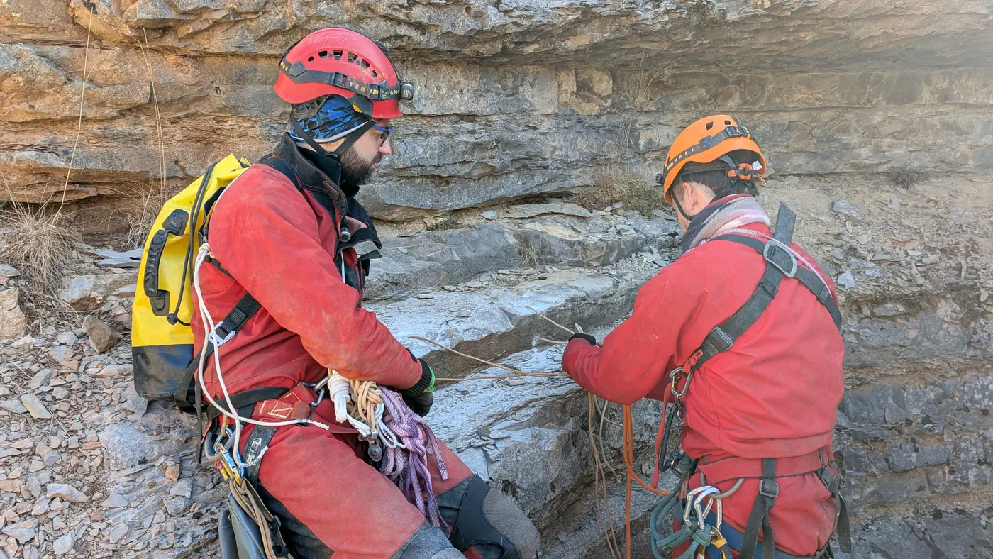 Deux spéléologues en combinaison rouge préparent un amarrage au pied d’une paroi rocheuse, en surface.