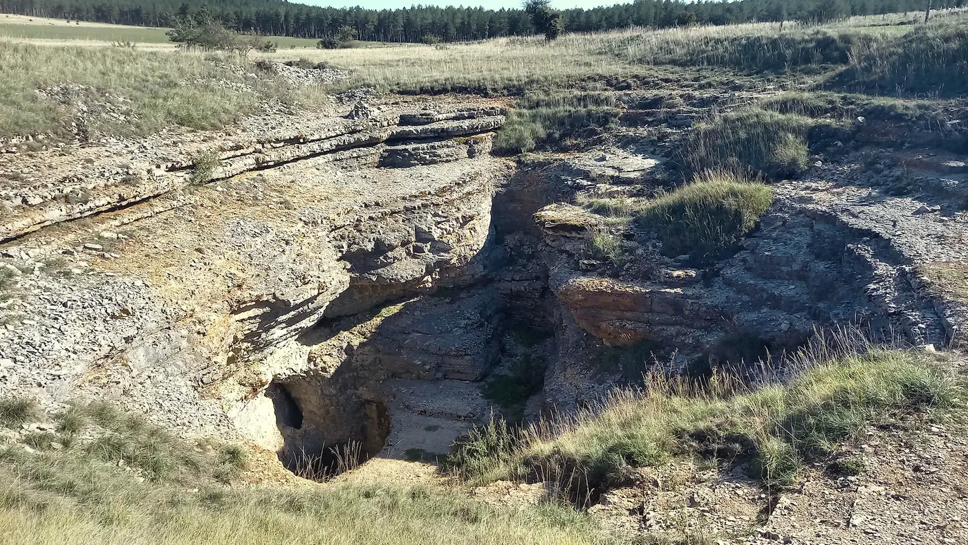 Sur le causse, une large dépression rocheuse aux strates calcaires s’ouvre sur l’entrée de la cavité.