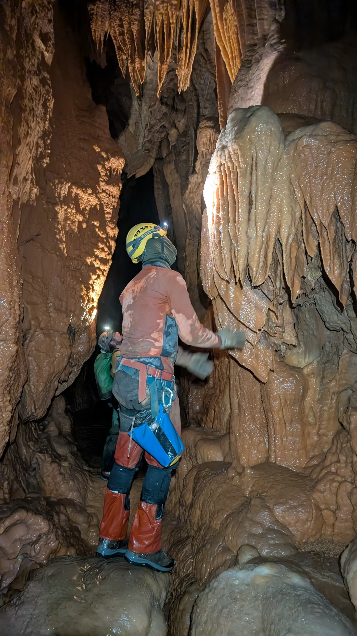 Un spéléologue en combinaison orange tend la main vers de grandes draperies de calcite ; un coéquipier le suit dans la galerie.